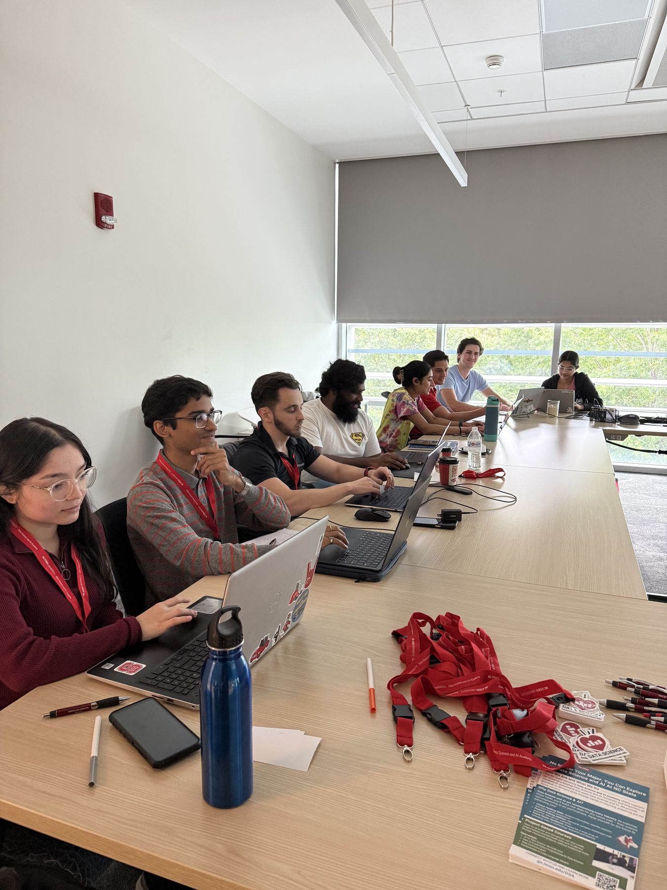 Participants wearing red lanyards work on laptops along a long conference table at the 2025 NC State hackathon
